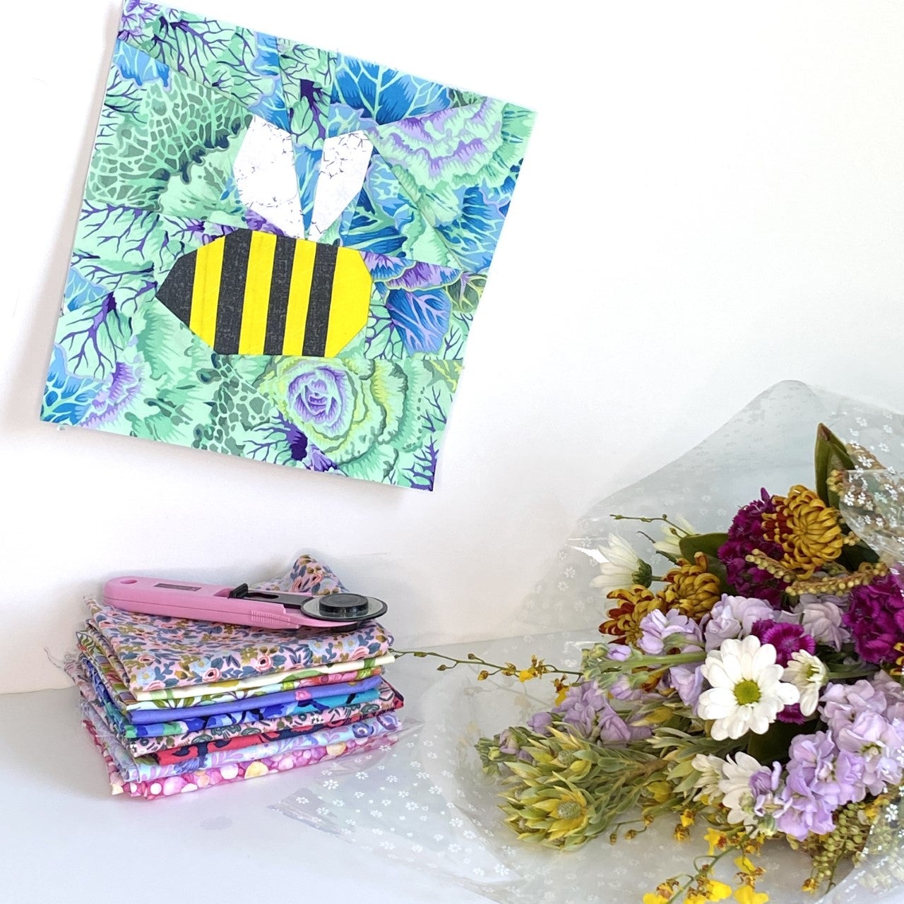 Bumble Bee Quilt Block Pattern. This sewing project is made using paper piecing and is super cute when combined with other quilt blocks such as flowers. The image shows a finished quilt block next to a stack of floral quilting fabrics and a rotary cutter, there is a bunch of flowers to the right of the bumble bee as if it is flying towards the flowers.