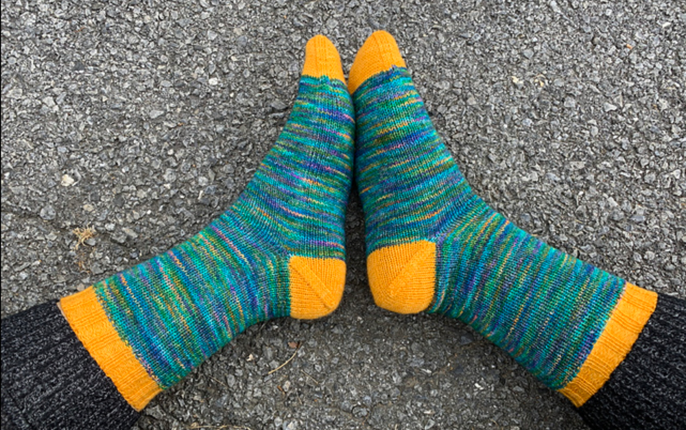 Tube socks in a variegated ocean-colored yarn. Heels, toes, and cuffs are bright yellow. They are modeled; the model is wearing black pants and the background is charcoal colored asphalt.